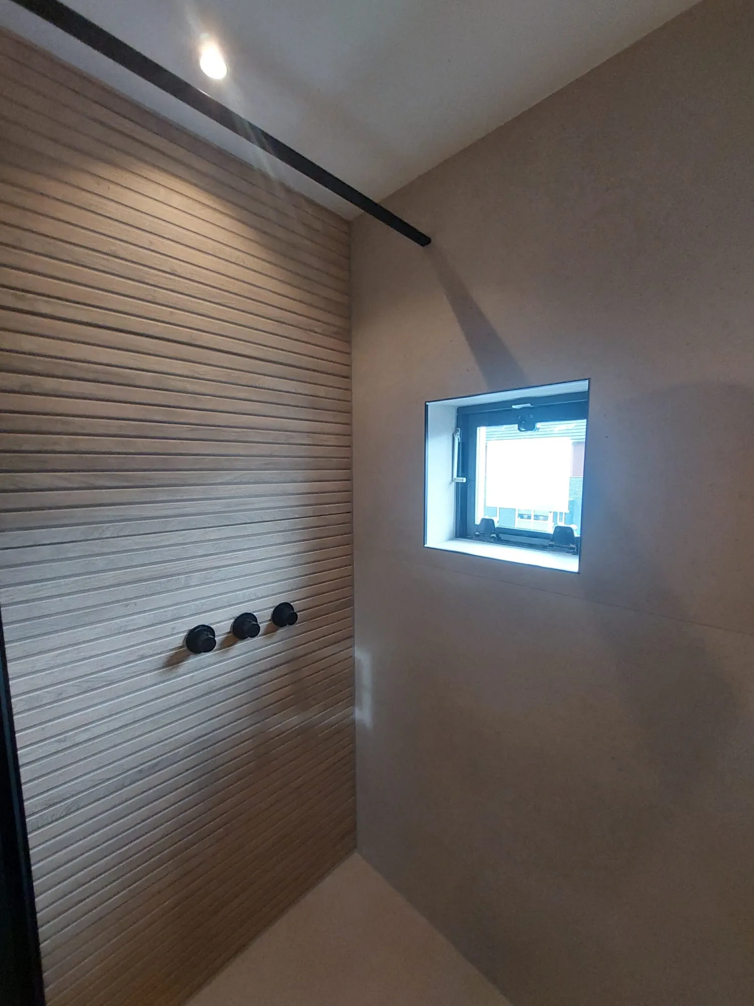 Minimalistische doucheruimte met glad stucwerk, horizontale houten lambrisering en zwarte bedieningsknoppen
