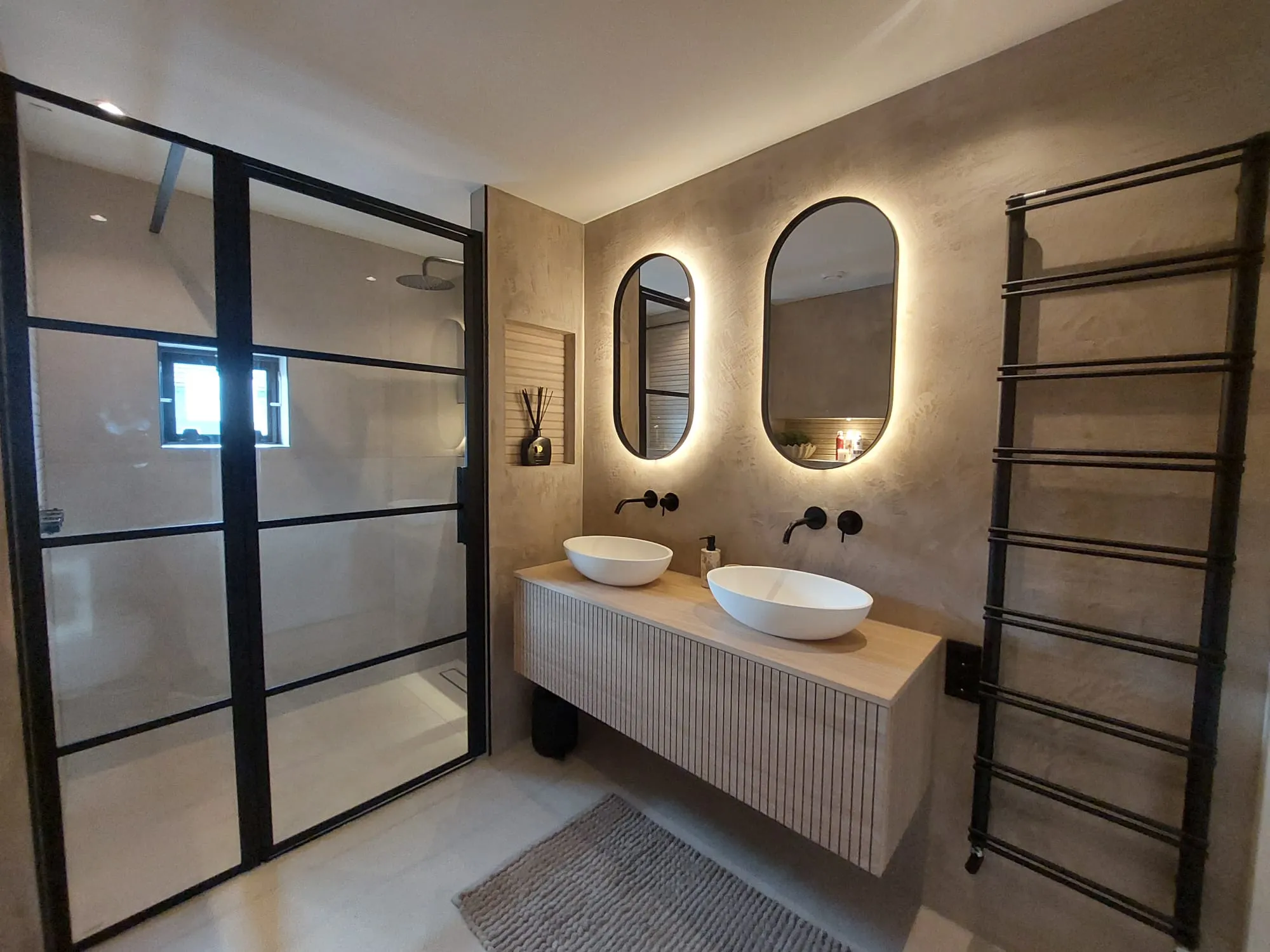 Moderne badkamer met stucwerk wanden, ovale verlichte spiegels, houten wastafelmeubel en zwart glazen douchecabine
