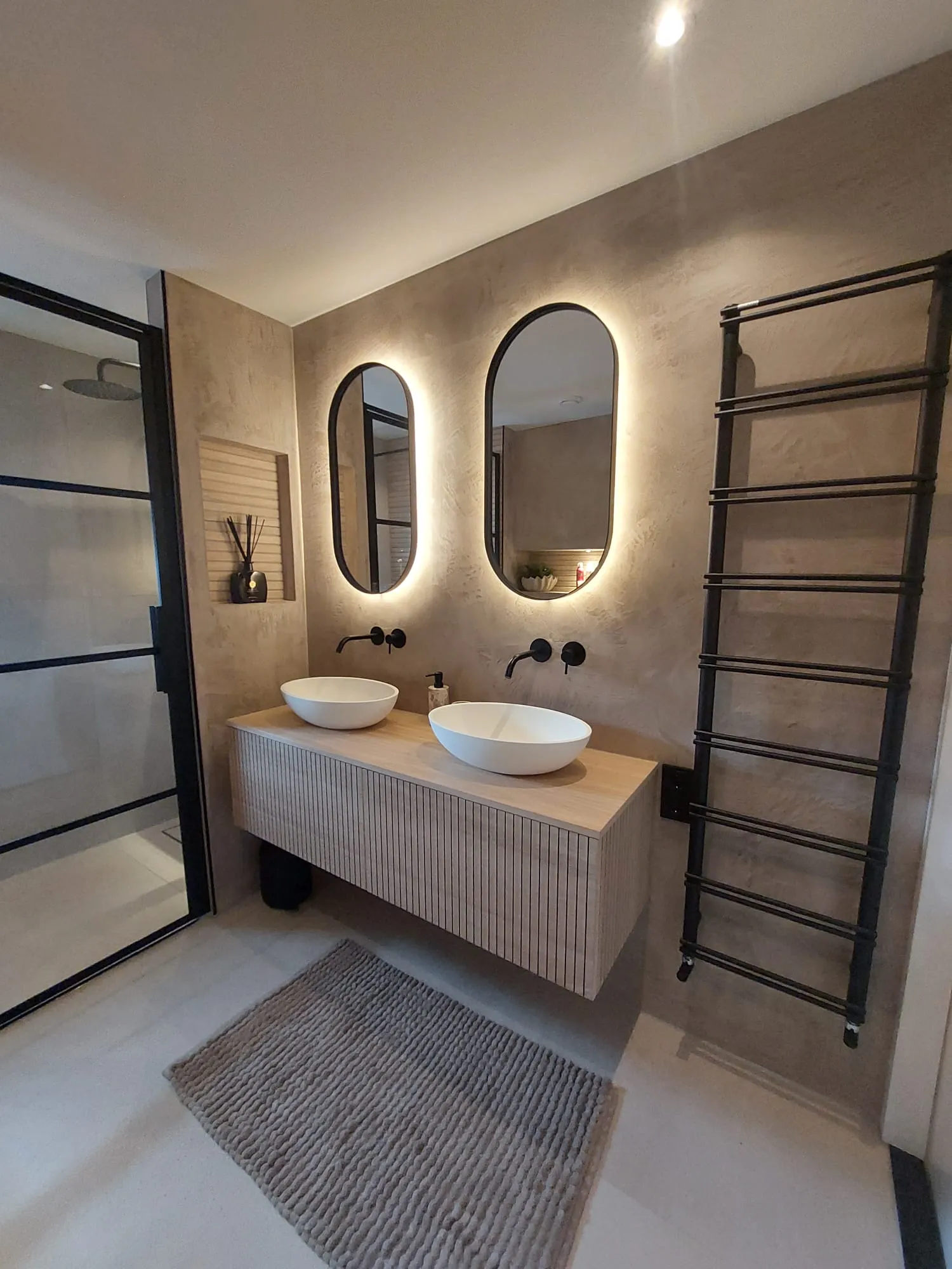 Moderne badkamer met stucwerk wanden, ovale verlichte spiegels, zwevend houten meubel met opzetwastafels
