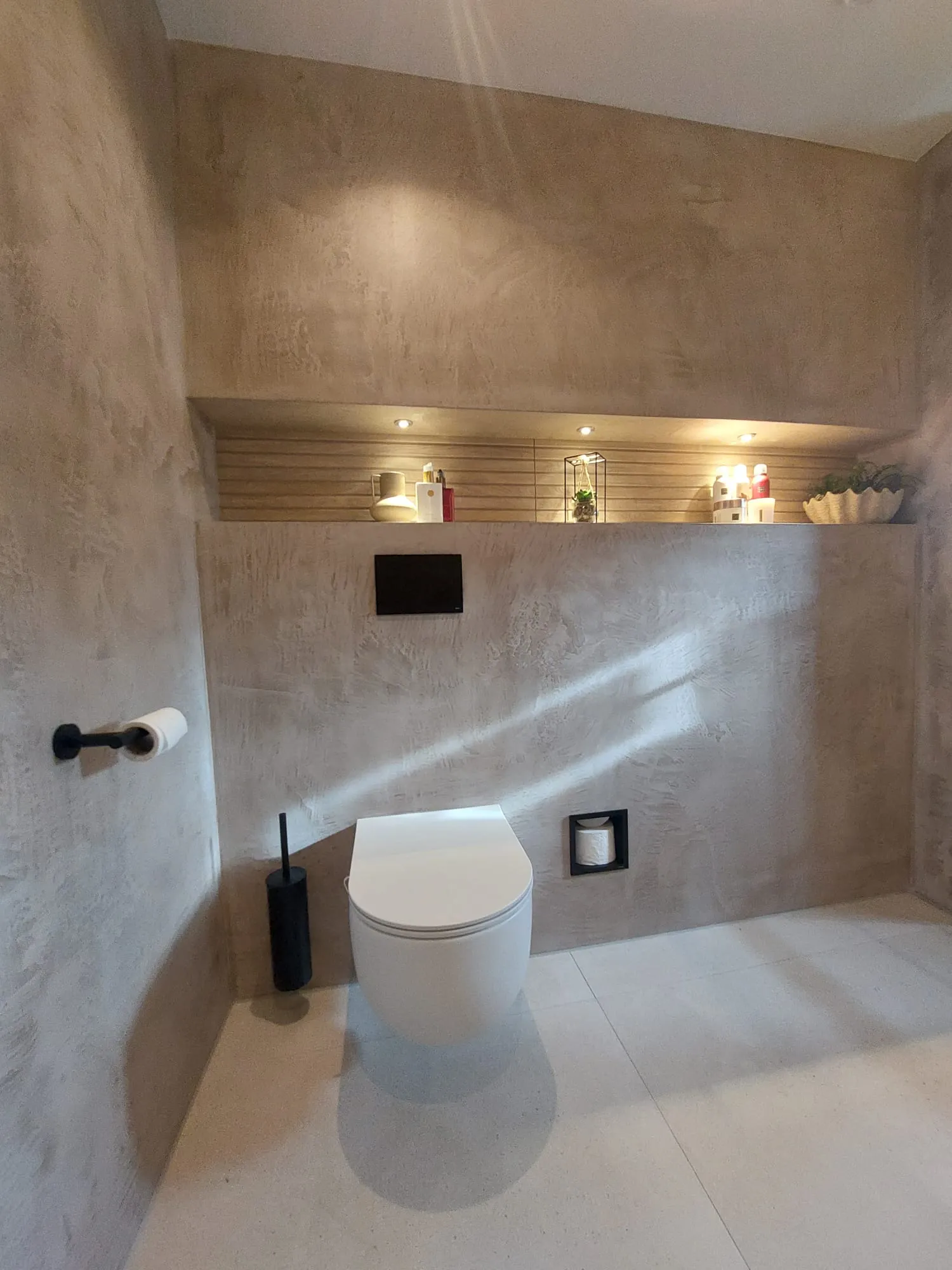 Minimalistisch toilet met glad stucwerk, wandgemonteerde WC en ingebouwde nis met toiletartikelen onder spotlights

