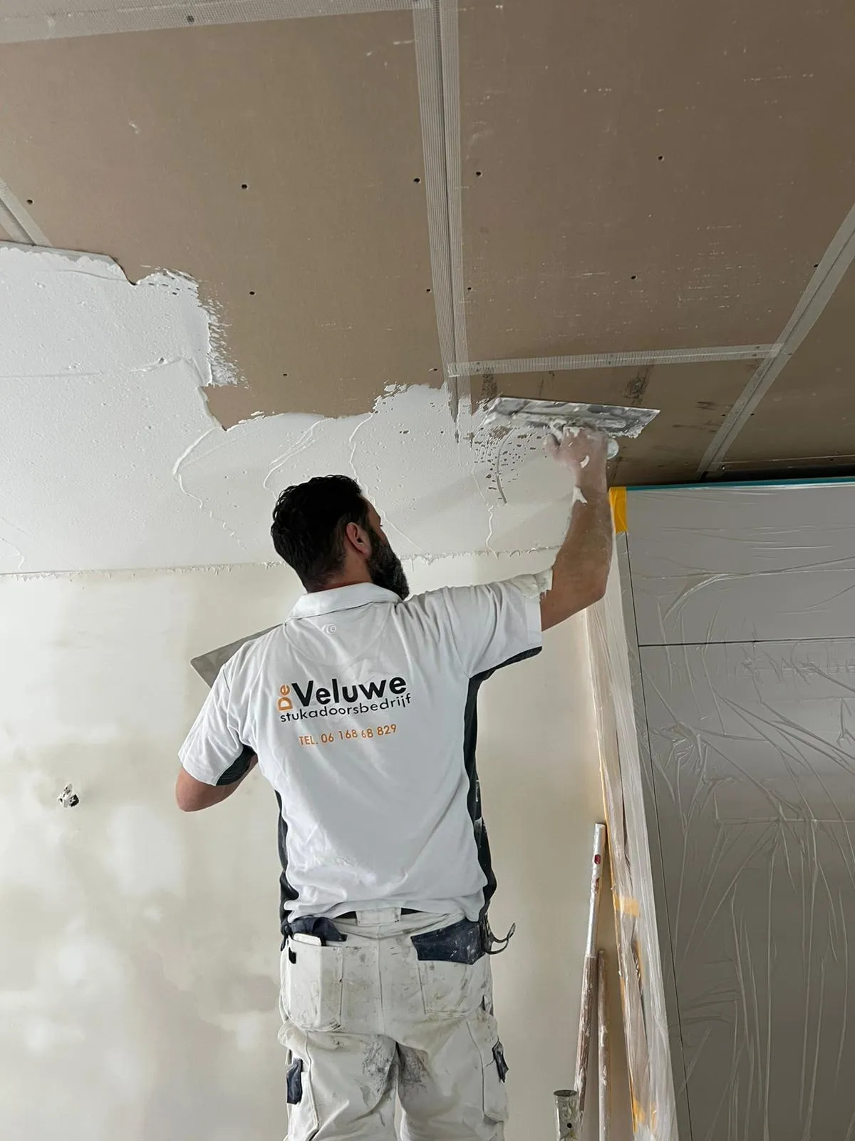 Professionele stukadoor van Stukadoorsbedrijf de Veluwe in witte werkkleding brengt stucwerk aan op plafond met truweel tijdens renovatie
