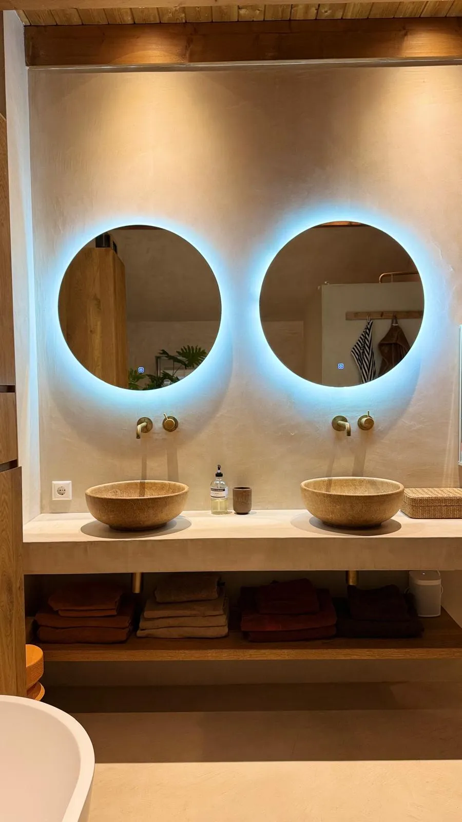 Moderne badkamer met glad stucwerk, ronde LED-verlichte spiegels, natuurstenen wastafels en messing kranen
