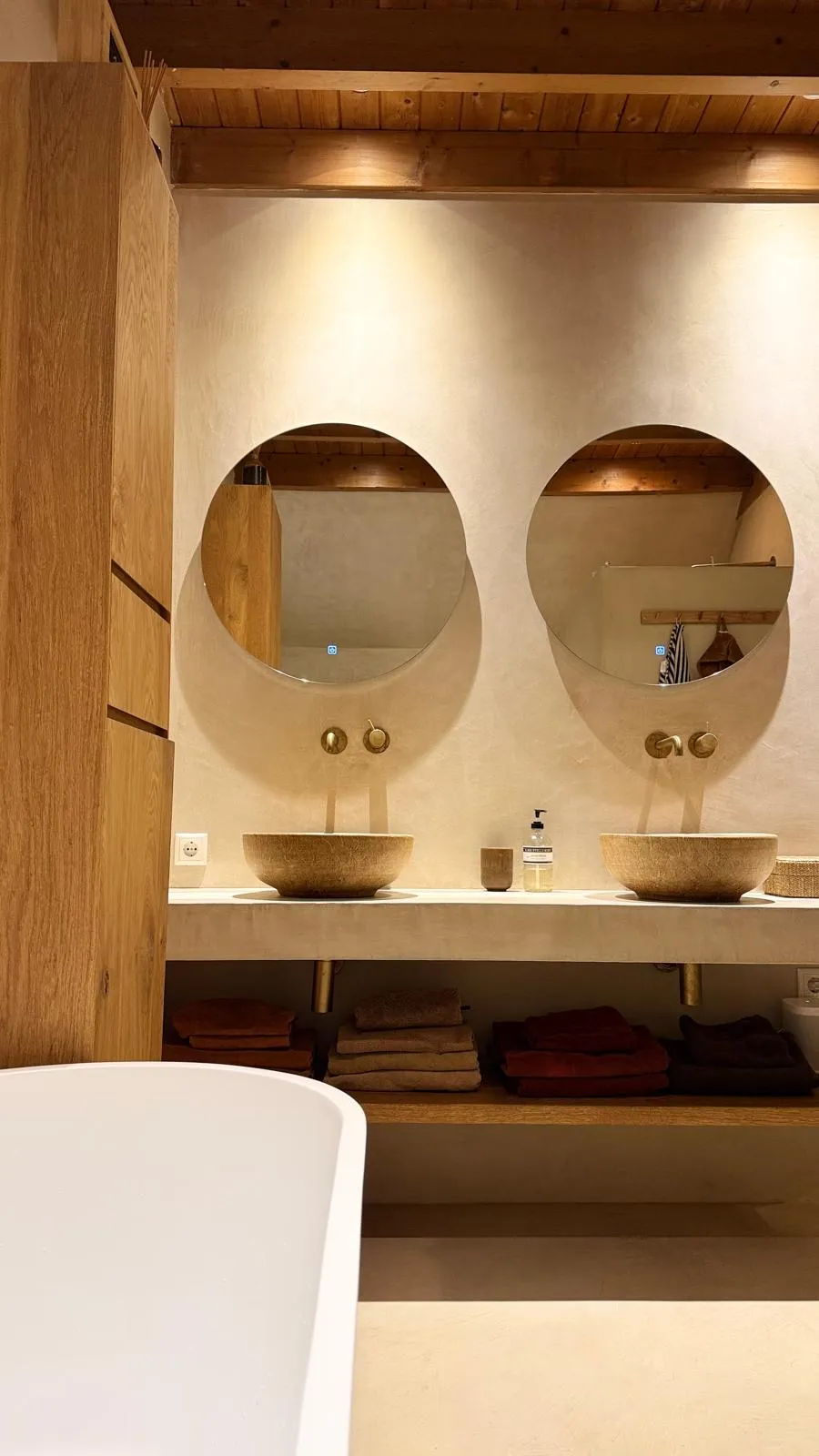 Moderne badkamer met stucwerk afwerking, houten plafond, natuurstenen wastafels en ronde spiegels
