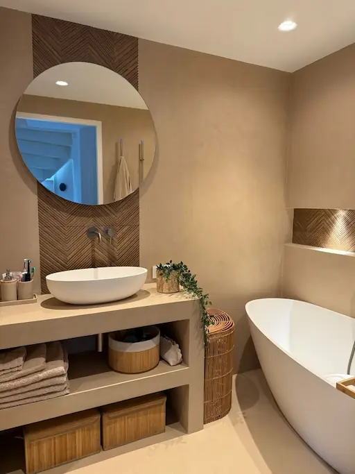 Moderne badkamer met glad stucwerk afwerking, beige blad met opzetwastafel, ronde spiegel en vrijstaand bad
