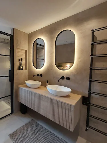 Moderne badkamer met stucwerk wanden, ovale verlichte spiegels, zwevend houten meubel met opzetwastafels
