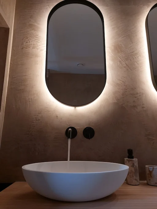 Moderne wastafel met glad stucwerk wandafwerking, ovale opzetkom, zwarte wandkraan en verlichte spiegel
