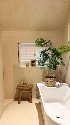 Minimalistische badkamer met beige decoratief stucwerk wandafwerking, vrijstaand bad met plant en houten krukje
