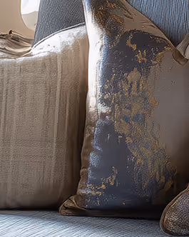 Close-up of two decorative pillows on a blue cushioned seat, one beige with a soft texture and the other metallic gray with a gold pattern.