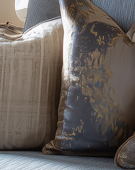 Close-up of two decorative pillows on a blue cushioned seat, one beige with a soft texture and the other metallic gray with a gold pattern.