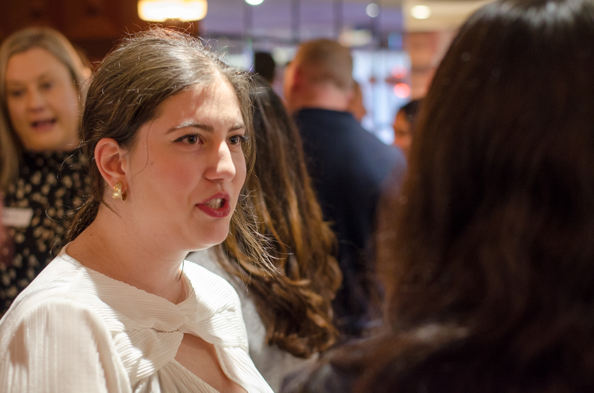 Young woman in a white blouse speaking to another person in a crowded indoor setting.