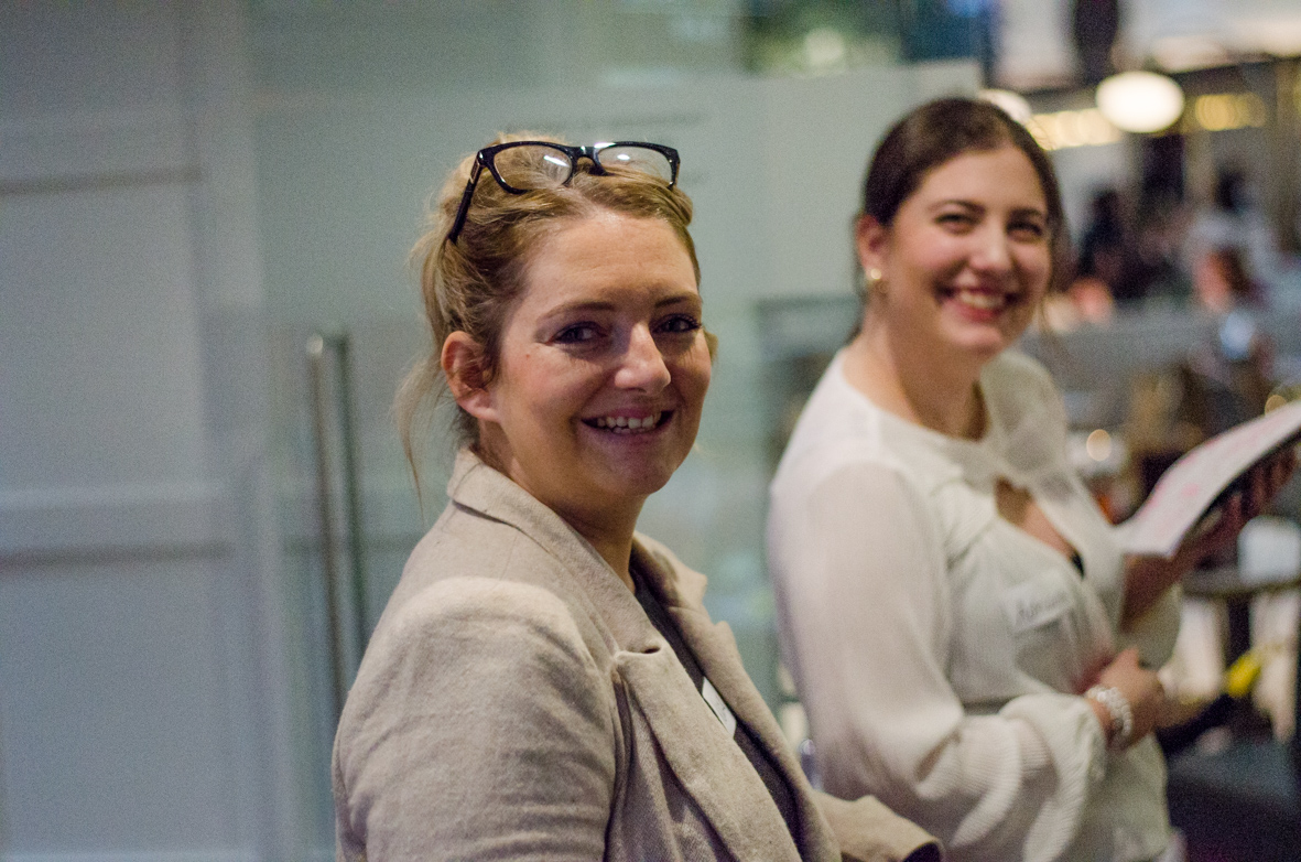 Two women smiling indoors, one with glasses on her head wearing a beige blazer and the other holding a document and wearing a white blouse.