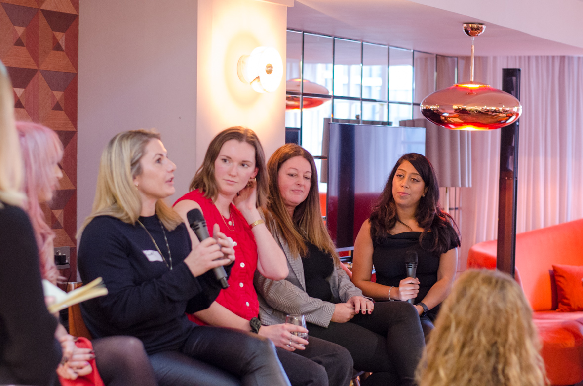 A group of women seated indoors, with one woman speaking into a microphone while others listen attentively.