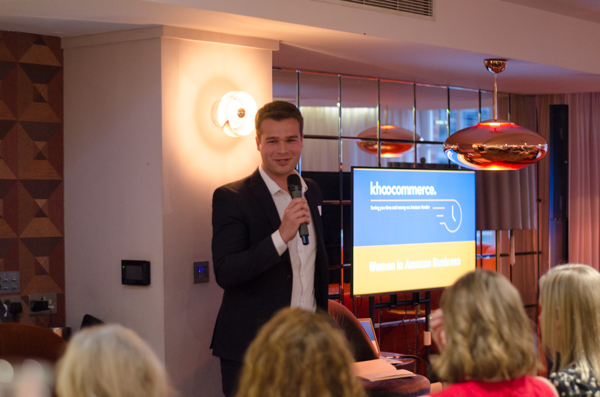 Man in a black suit holding a microphone speaking to an audience with a screen behind him displaying 'khoocommerce' and 'Women in Amazon Business'.
