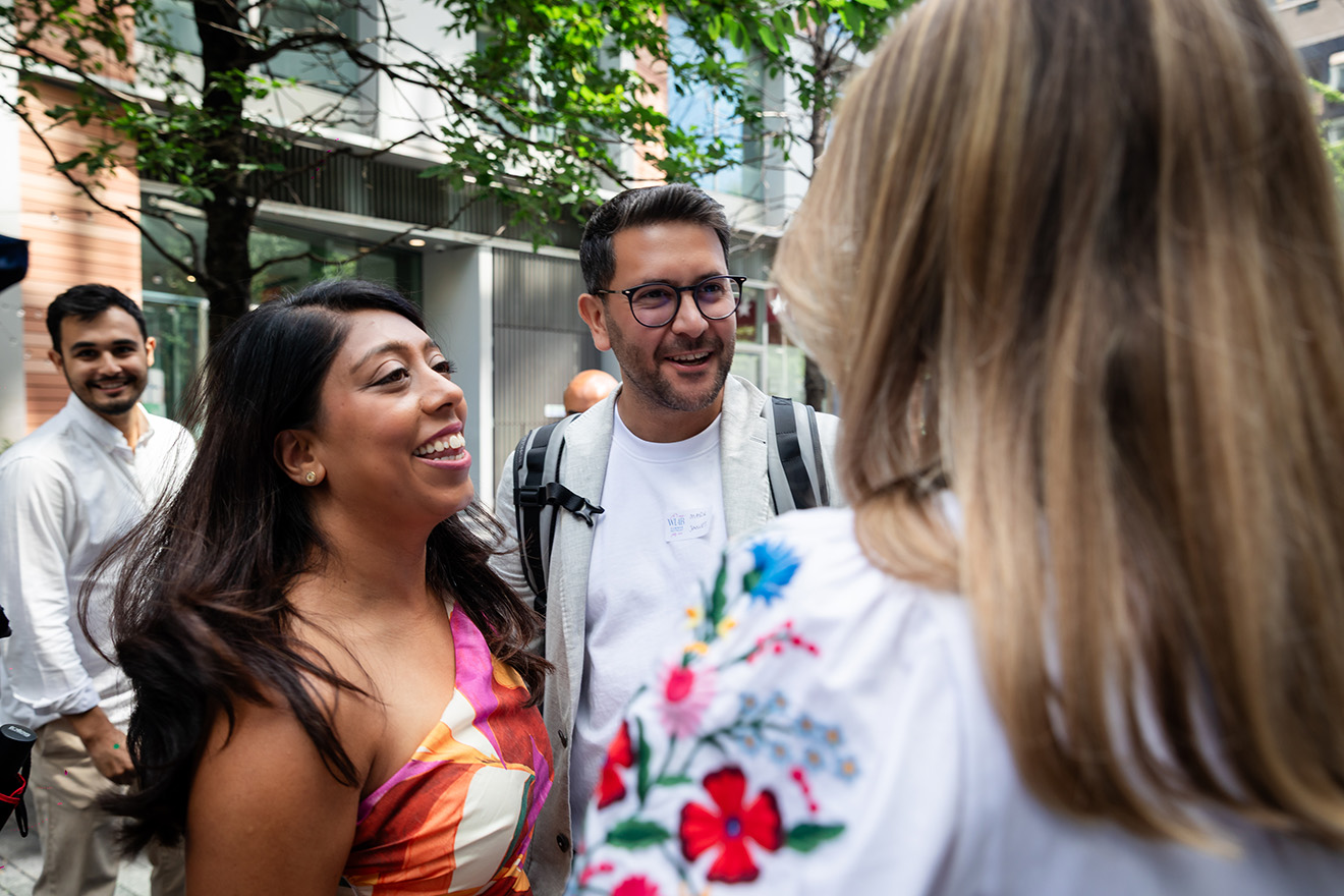 Two women smiling indoors, one wearing glasses on her head and a beige jacket, the other holding a paper and wearing a white blouse.