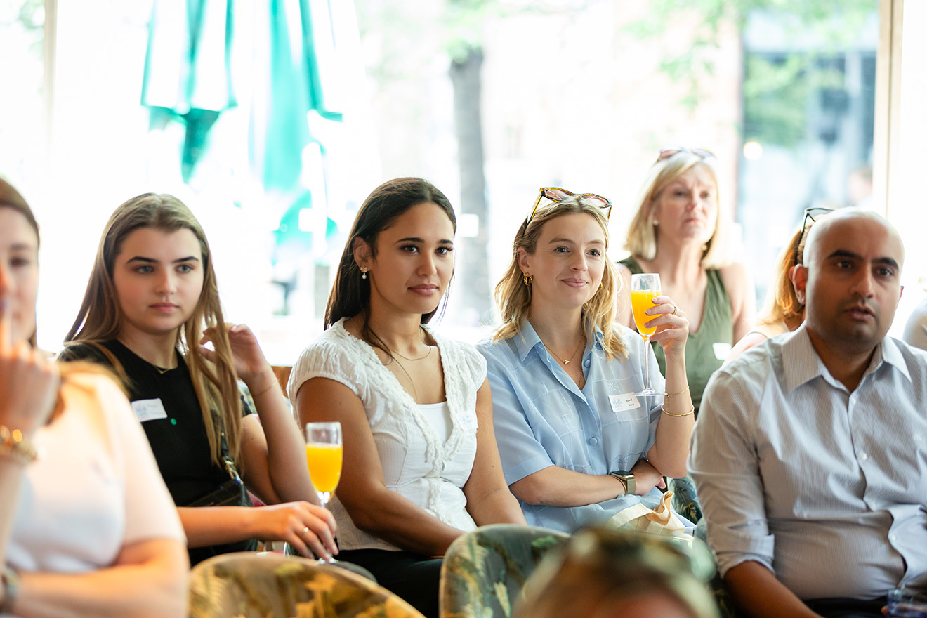 Two women smiling indoors, one wearing glasses on her head and a beige jacket, the other holding a paper.