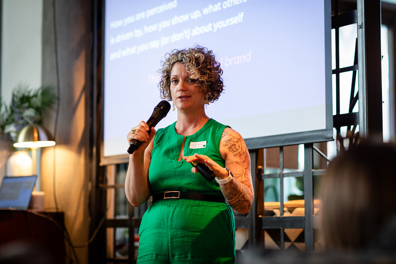 Man in suit holding a microphone and speaking in front of an audience with a screen displaying 'khoocommerce' and 'Women in Amazon Business'.
