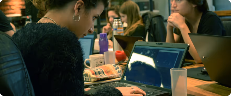 Group of people working on laptops around a shared table in a collaborative workspace, with a close-up of one person focused on their screen.