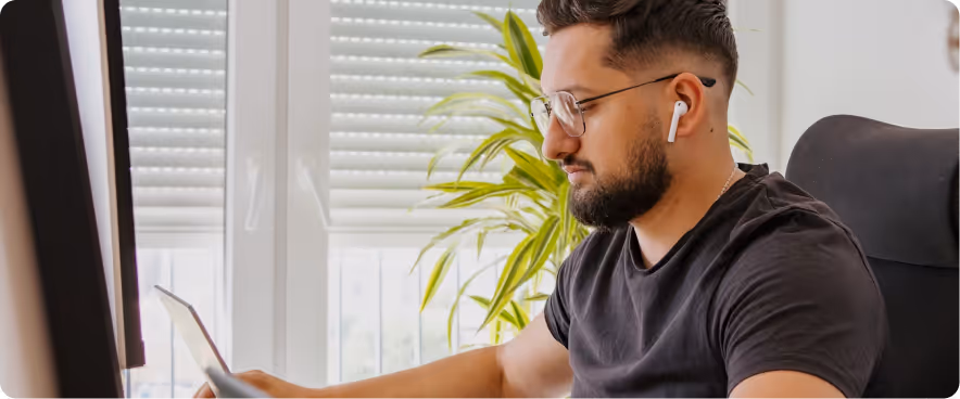 Man wearing wireless earbuds and glasses working on a laptop at home, sitting near a bright window and indoor plant.