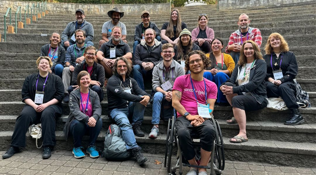 Group of 21 people sitting on outdoor stone steps smiling at the camera, including one person in a wheelchair wearing a bright pink shirt.