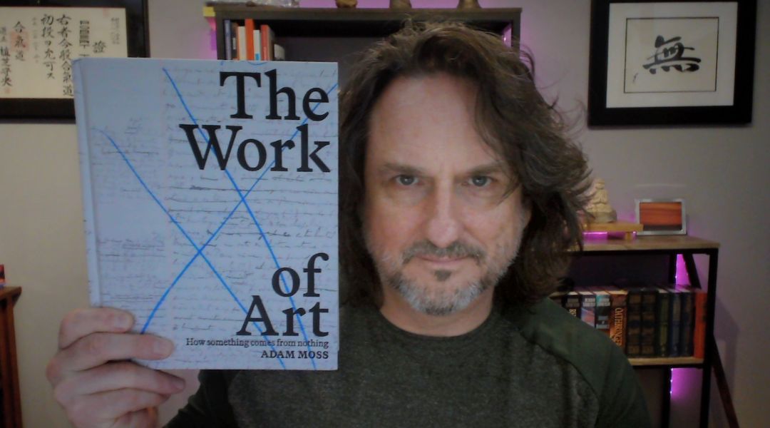 Man with shoulder-length hair holding a book titled 'The Work of Art' by Adam Moss.