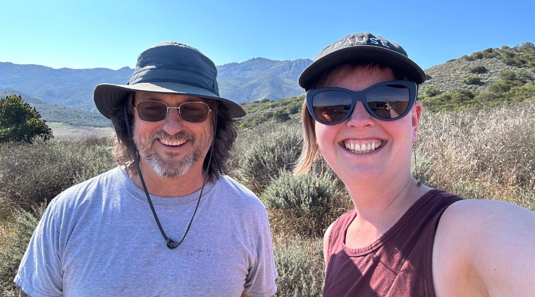 Two people smiling outdoors in a sunny mountainous area with shrubs and clear blue sky.
