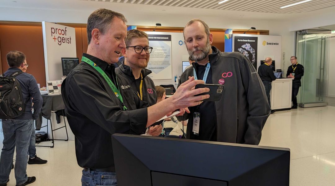 Three men standing in a conference area, one showing something on a smartphone to the other two.