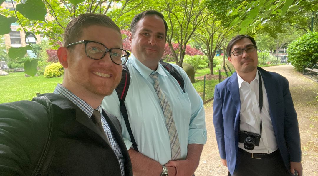 Three men in business attire posing for a selfie outdoors in a green park with trees and a walking path.