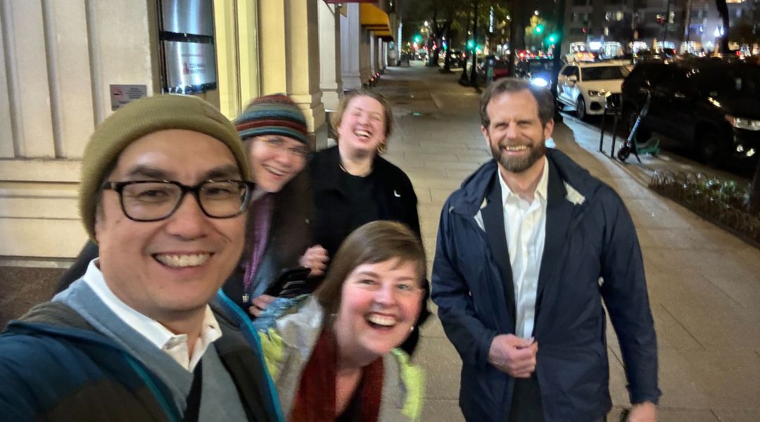 Group of five people smiling and laughing on a city sidewalk at night with streetlights and cars in the background.