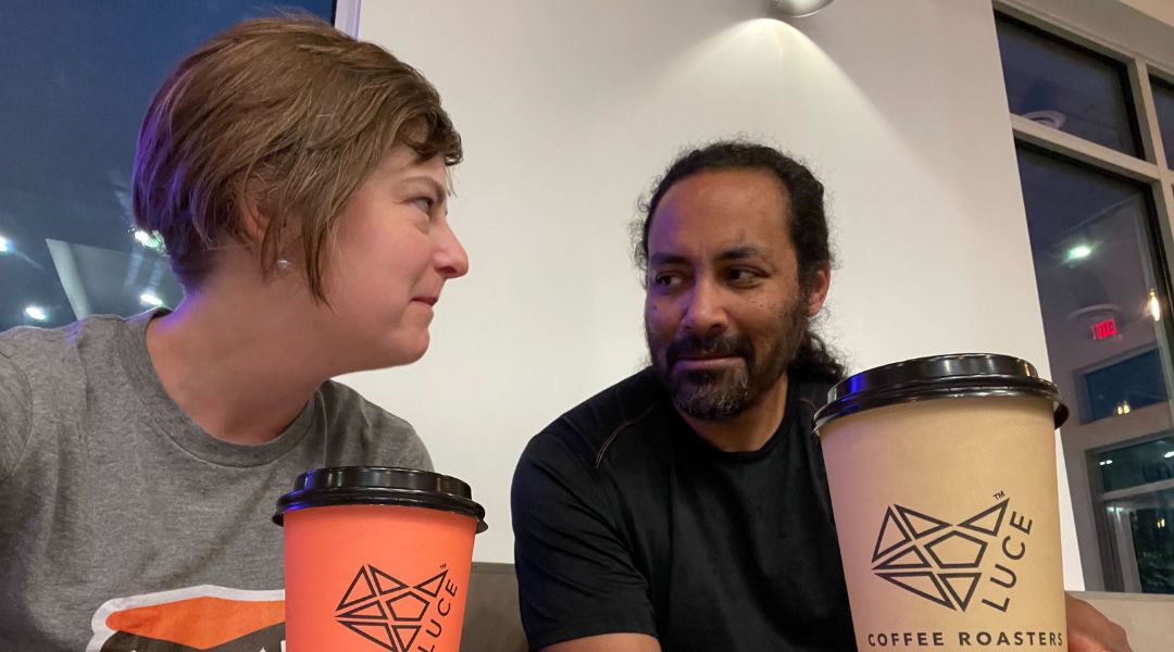 A woman and a man sitting side by side indoors, each holding a coffee cup from Luce Coffee Roasters, looking at each other.