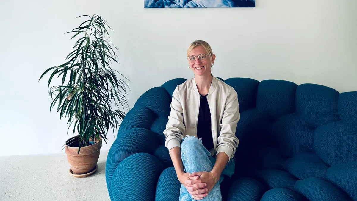 Smiling woman with glasses wearing a light jacket and jeans sitting on a blue bubble sofa next to a potted plant.