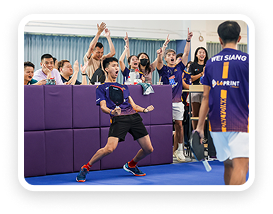Young boy in a sports uniform celebrating a point during a game, with an enthusiastic crowd cheering behind a purple barrier.