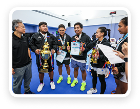 Four athletes wearing medals holding trophies and certificates, standing in a sports hall with two officials.