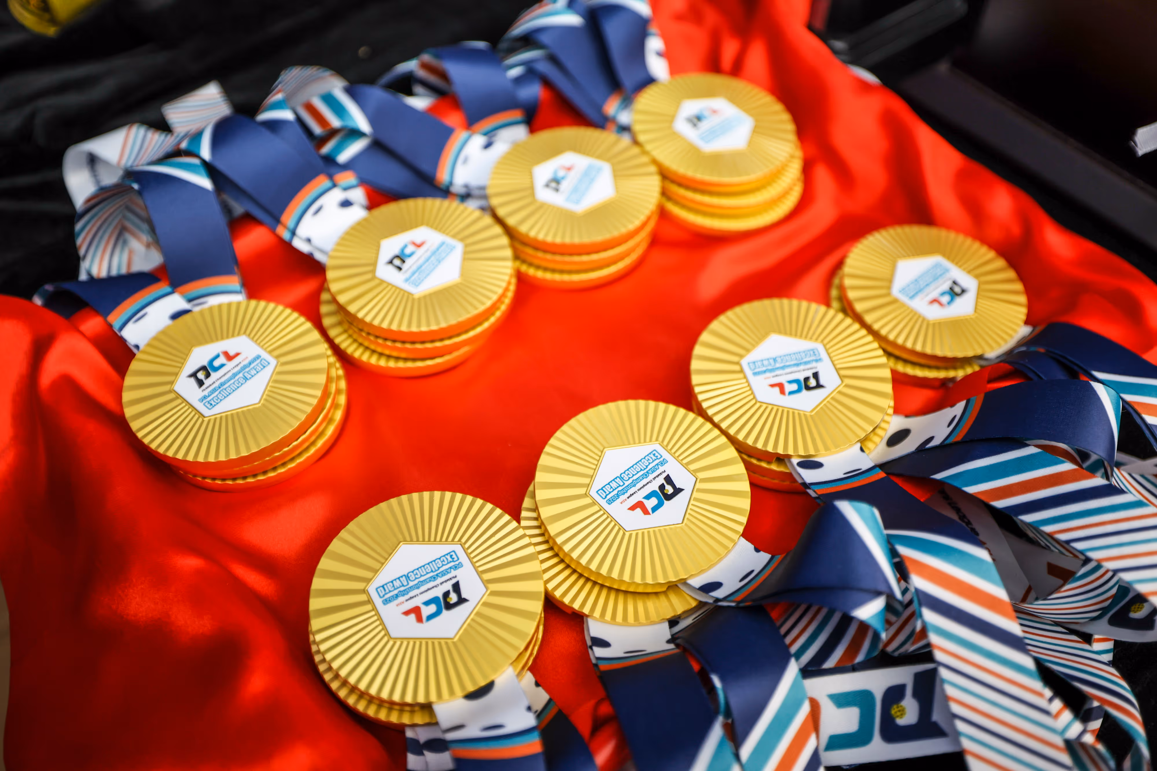 Multiple stacks of gold round medals with blue, white, and orange striped ribbons placed on red fabric.