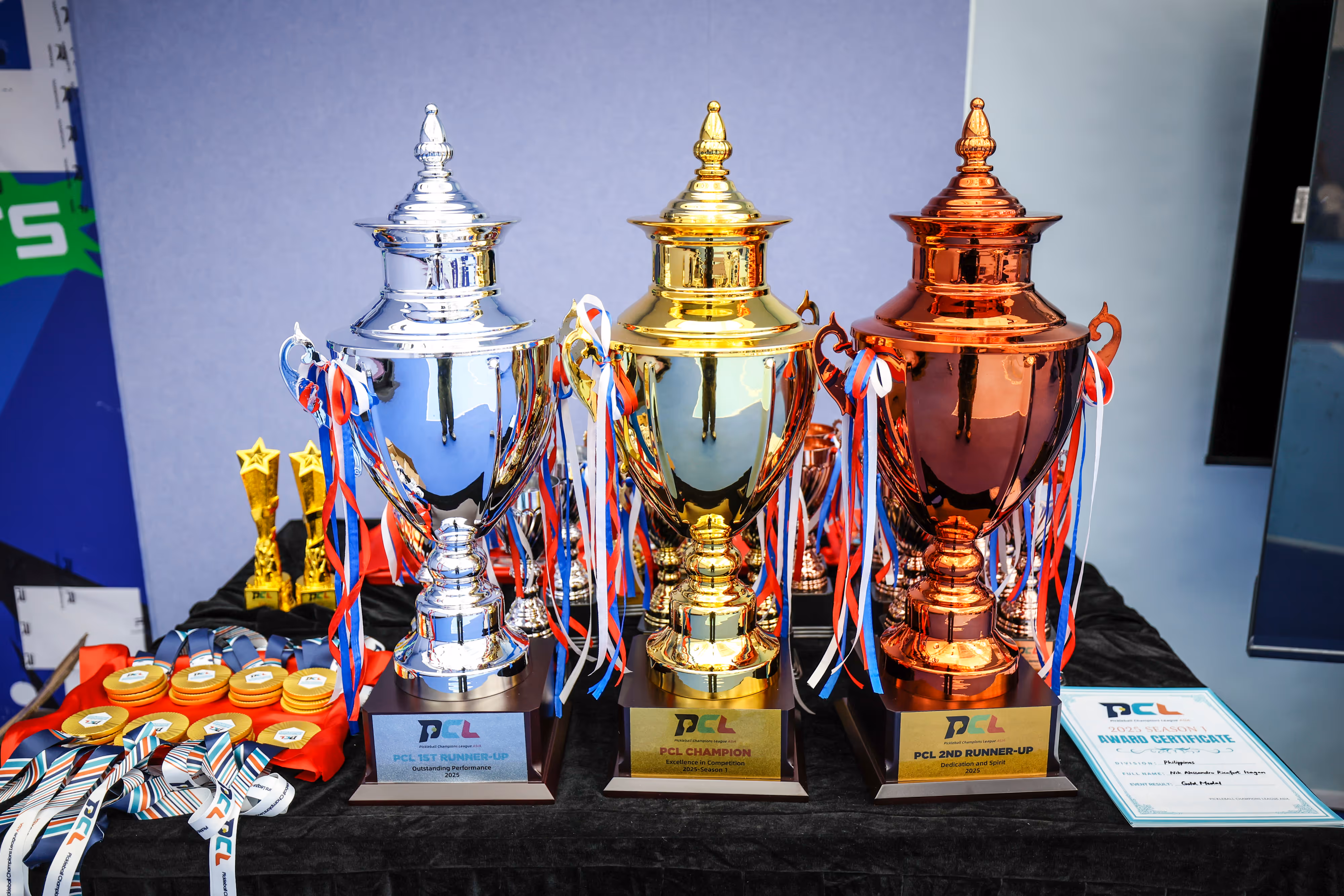 Three large trophies in silver, gold, and bronze with red, white, and blue ribbons, surrounded by medals and a certificate on a black table.