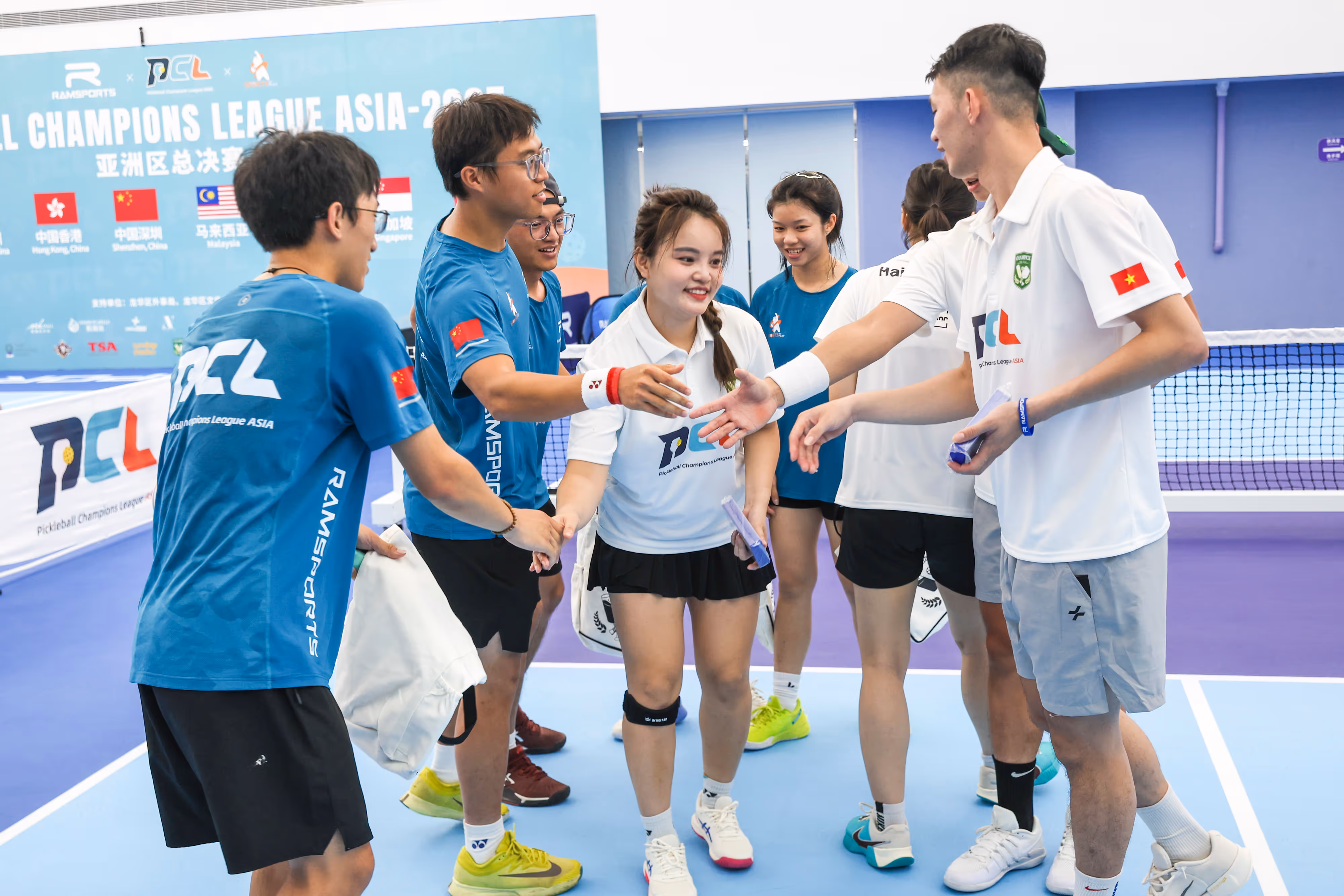 Two mixed teams in blue and white jerseys shaking hands on an indoor pickleball court at the Pickleball Champions League Asia event.