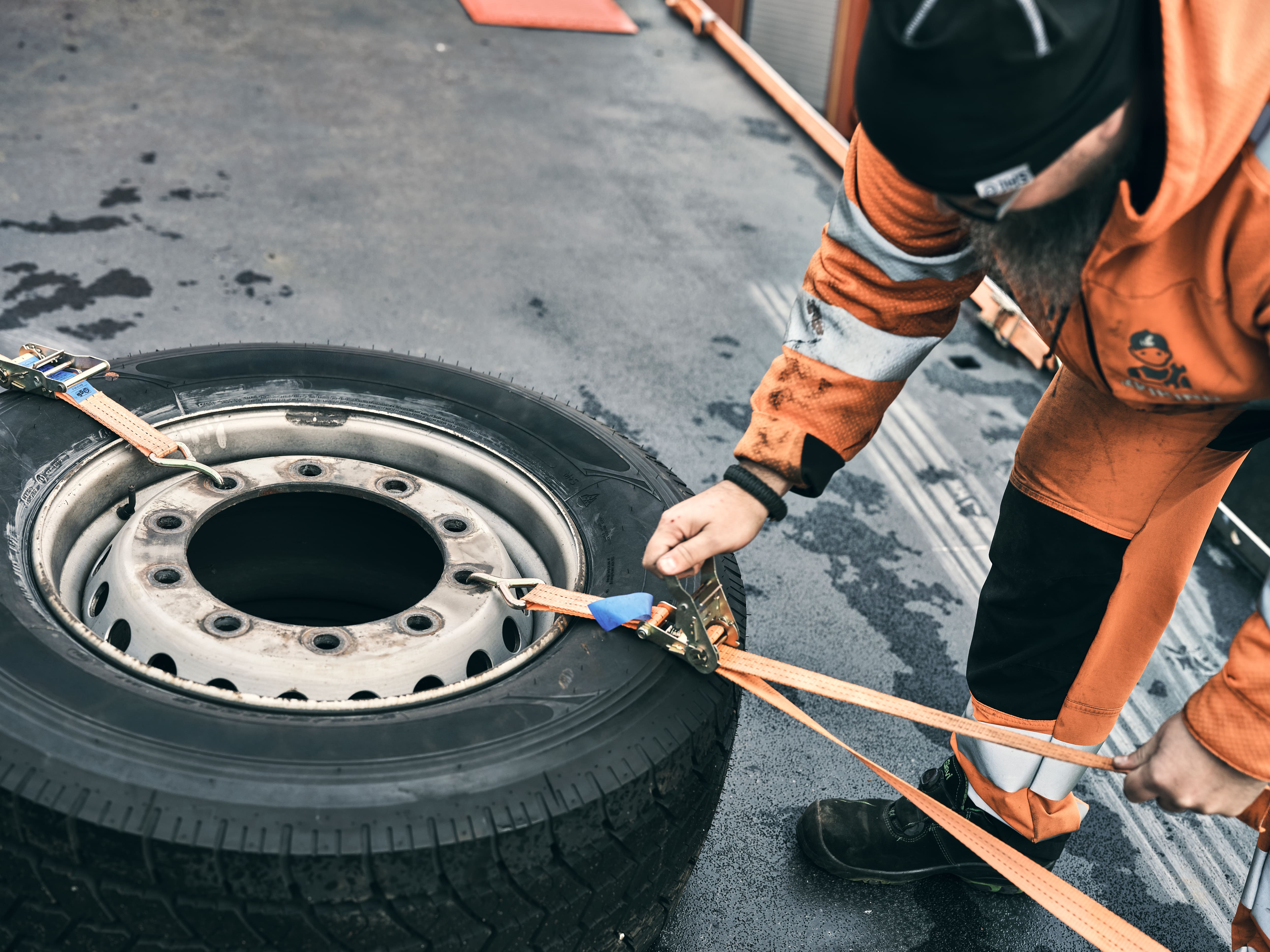 Worker in orange safety clothing securing a large tire with orange ratchet straps on a wet surface.