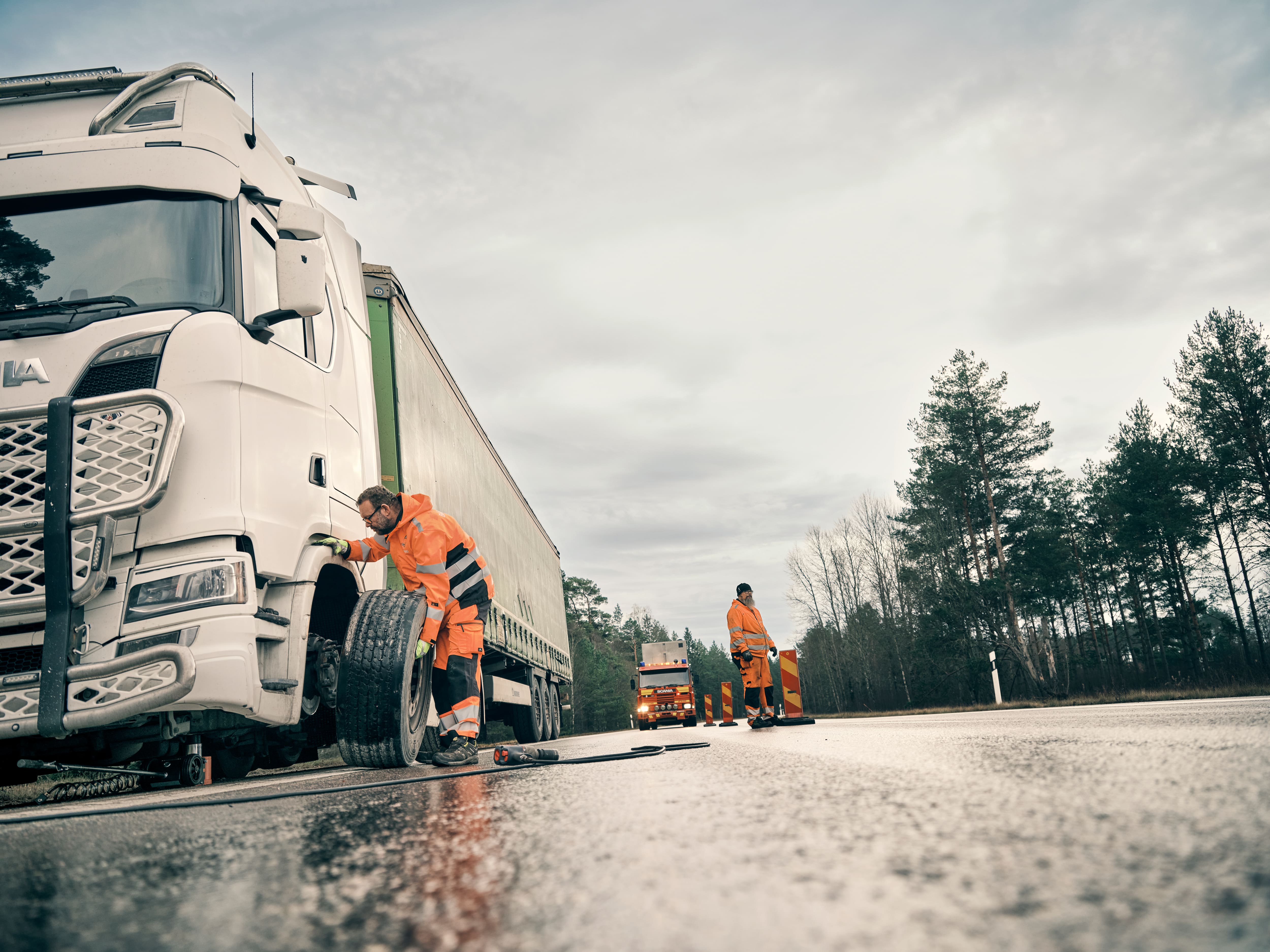 Two men in orange safety gear changing a tire on a white semi-truck on a wet road with traffic cones and a service truck in the background.