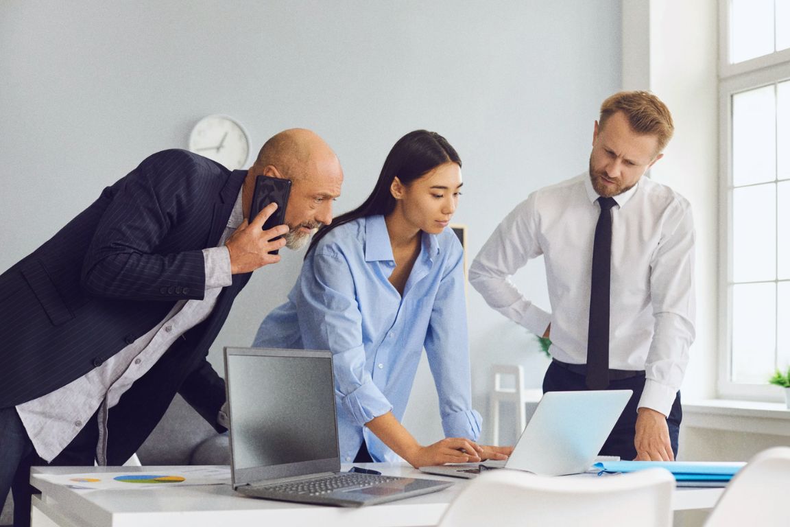 Three business professionals collaborating in an office, with one man on the phone and two others focused on a laptop.