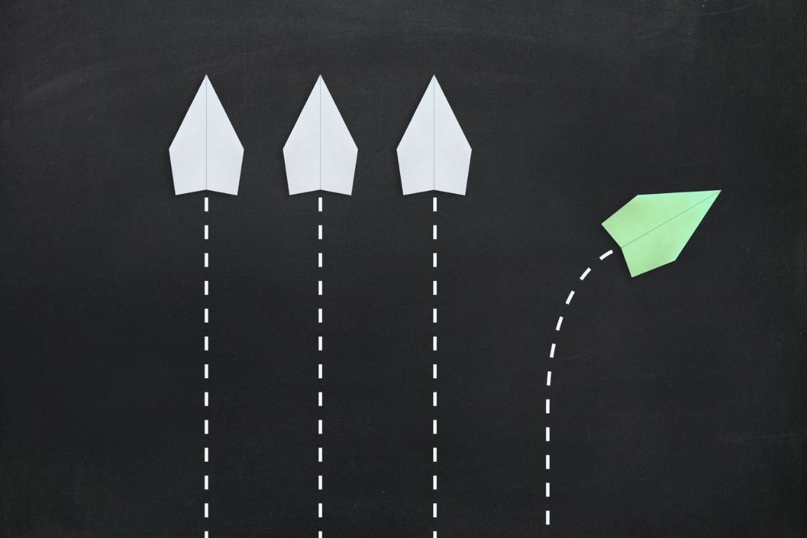 Three white paper airplanes flying straight with dashed lines and one green paper airplane diverging to the right on a black background.