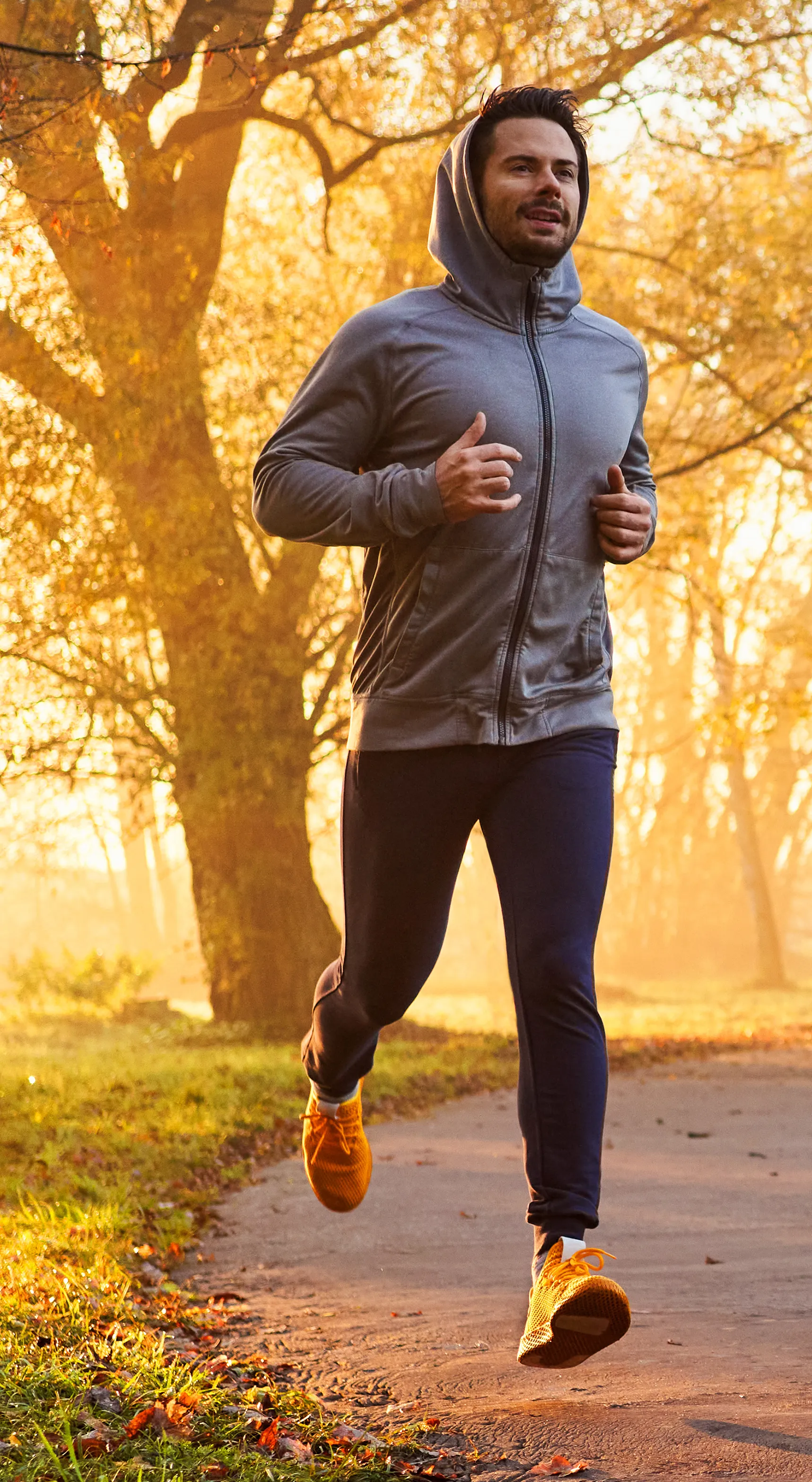 Man jogging in a park wearing a gray hoodie and orange sneakers, surrounded by autumn leaves and morning sunlight, promoting health and wellness related to erectile dysfunction treatments.