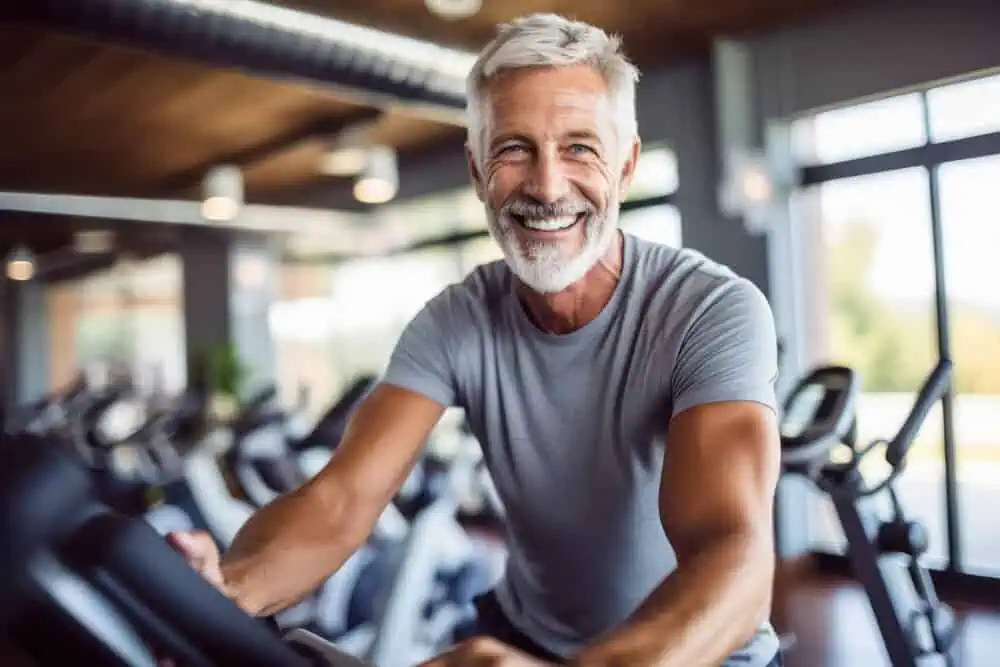 Smiling older man exercising on a stationary bike in a fitness center, highlighting health and vitality related to effective erectile dysfunction treatments.