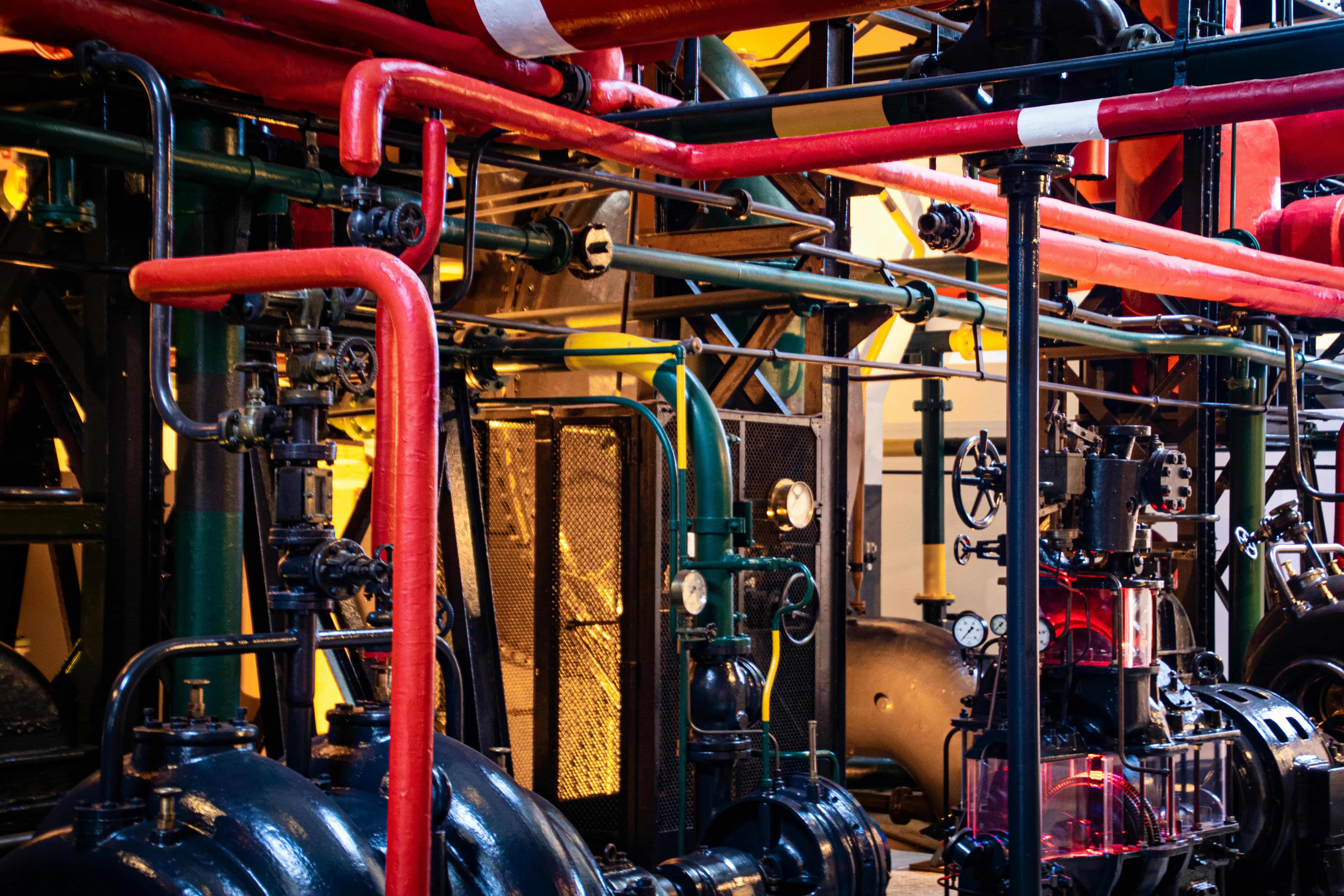 A complex network of colorful industrial pipes and valves in a machinery-filled factory setting.