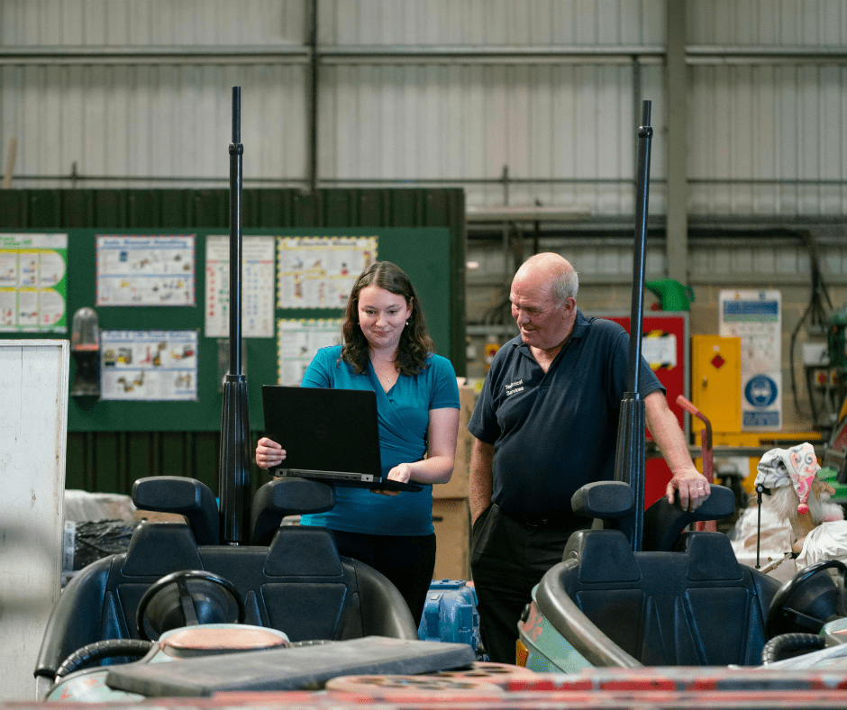A woman in a blue polo shirt and a man in a work uniform reviewing machinery data on a laptop in a manufacturing plant.