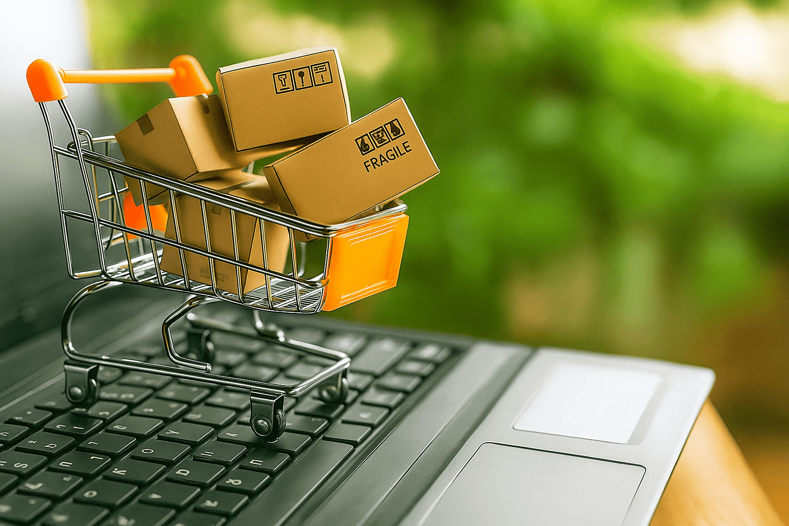 A miniature shopping cart filled with small cardboard boxes sits on a laptop keyboard, symbolizing online shopping and e-commerce, with a green blurred background.