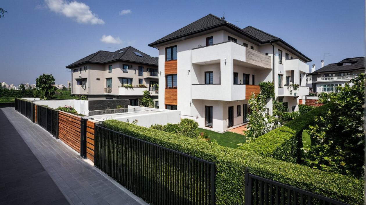Modern fehér, többszintes házak sötét tetővel és fa díszítéssel, zöld sövényekkel és fekete kerítéssel körülvéve, kék ég alatt.