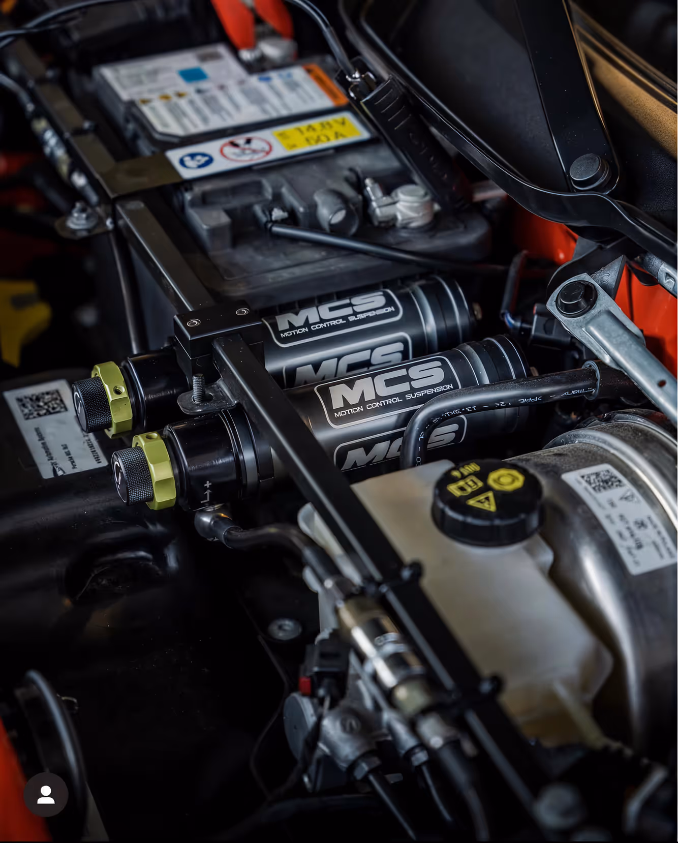 Close-up of car engine components featuring dual MCS Motion Control Suspension canisters and various hoses and connectors.