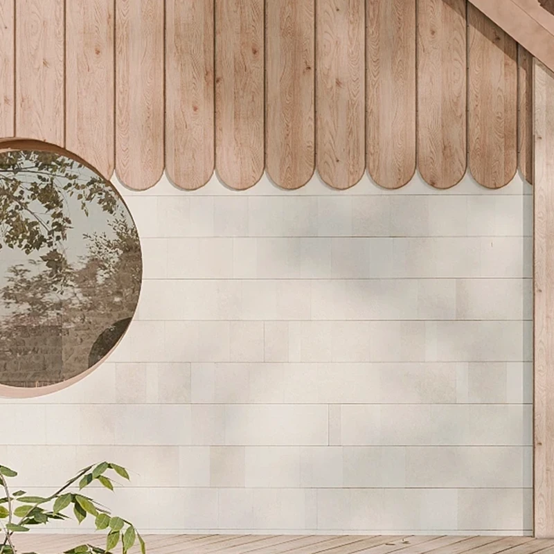 Exterior wall with light wood scalloped siding at the top and white horizontal stone tiles below with a round window reflecting tree branches.