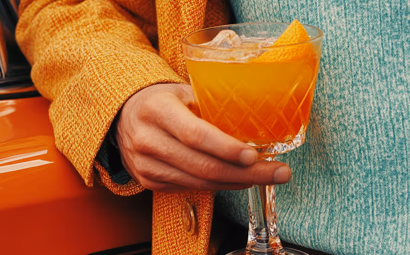 Person holding a vintage glass with an orange cocktail, garnished with an orange slice, wearing an orange textured coat and a green knit sweater.