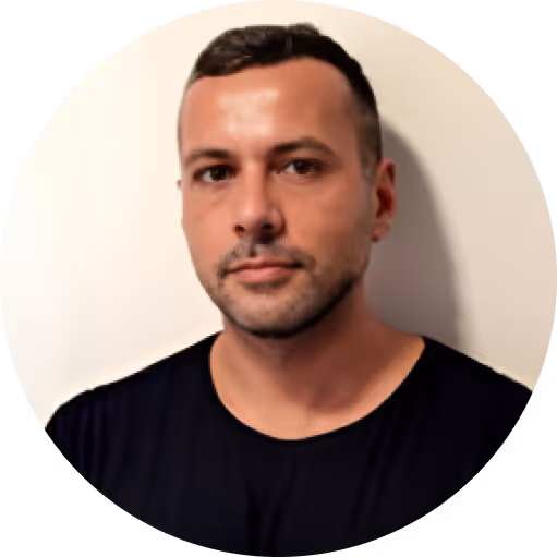 Portrait of a man with short dark hair and stubble wearing a black shirt against a light background.