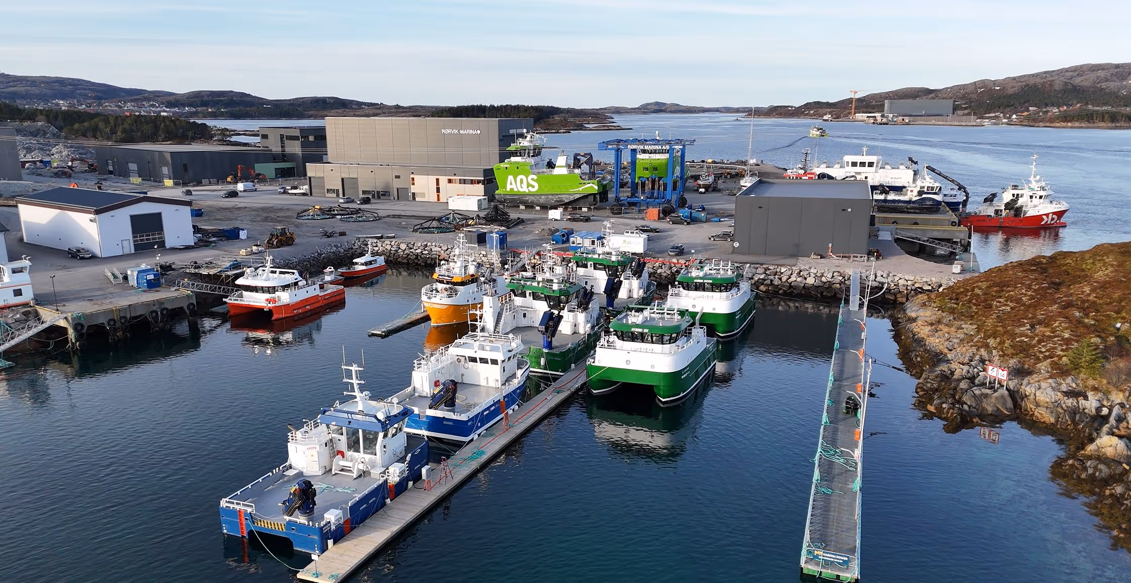 Havn med flere små fiskebåter og båter fortøyd til brygge, omgitt av industri- og lagerbygninger i et kystlandskap.